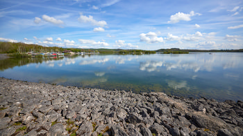 This landscape photograph captures Carsington Water harbour during a spring morning, located in the Peak District. The main subject of the image is the clear reflection of boats moored at the edge of the reservoir, visible across the calm surface of the lake. The tranquil water creates a mirror effect, displaying both the boats and the surrounding greenery. Rocky shorelines are prominent in the foreground, while the background features wooded areas and gently rolling hills, characteristic of the Peak District. The overall scene demonstrates the serenity and clarity of a spring morning at Carsington Water, with notable reflections enhancing the sense of calm across the lake’s surface.